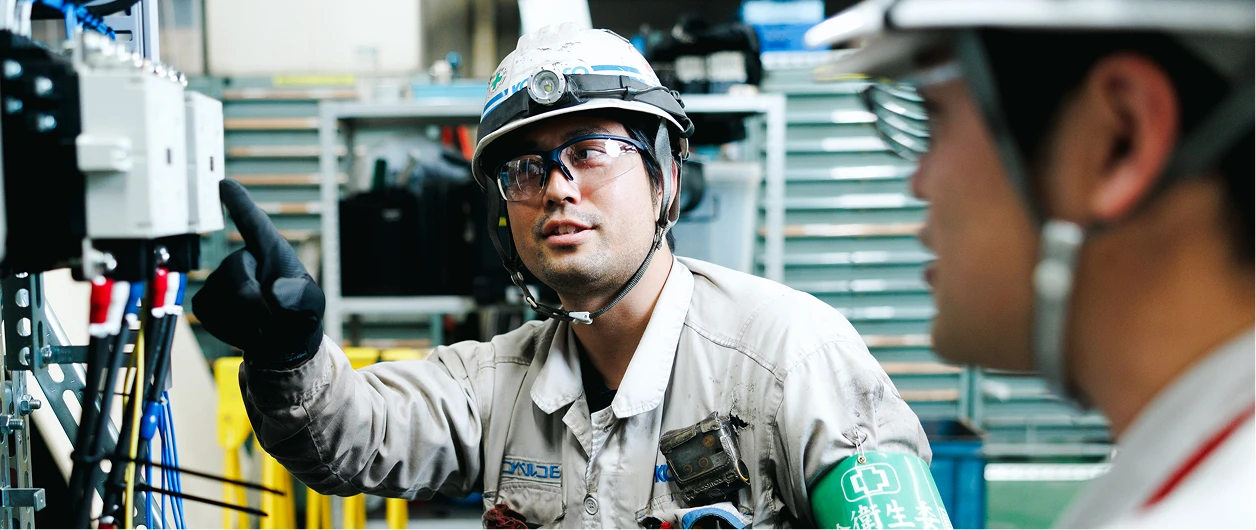 工場内で配電盤の修理を社員に説明する工事監督職の写真