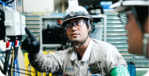 工場内で配電盤の修理を社員に説明する工事監督職の写真